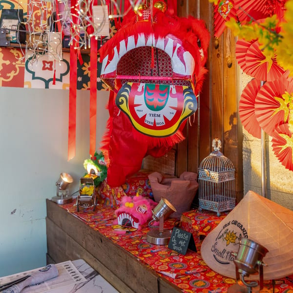 Vietnamese decor with a red lion dance head, conical hat, and red patterned fabric on a wooden counter.