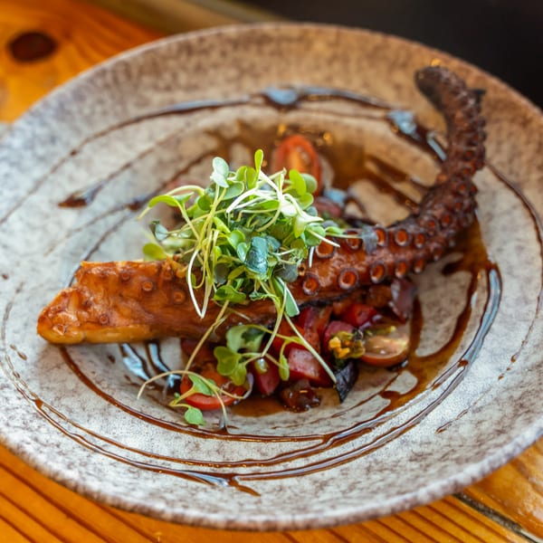 Grilled octopus with microgreens, cherry tomatoes, and a balsamic glaze on a speckled plate.